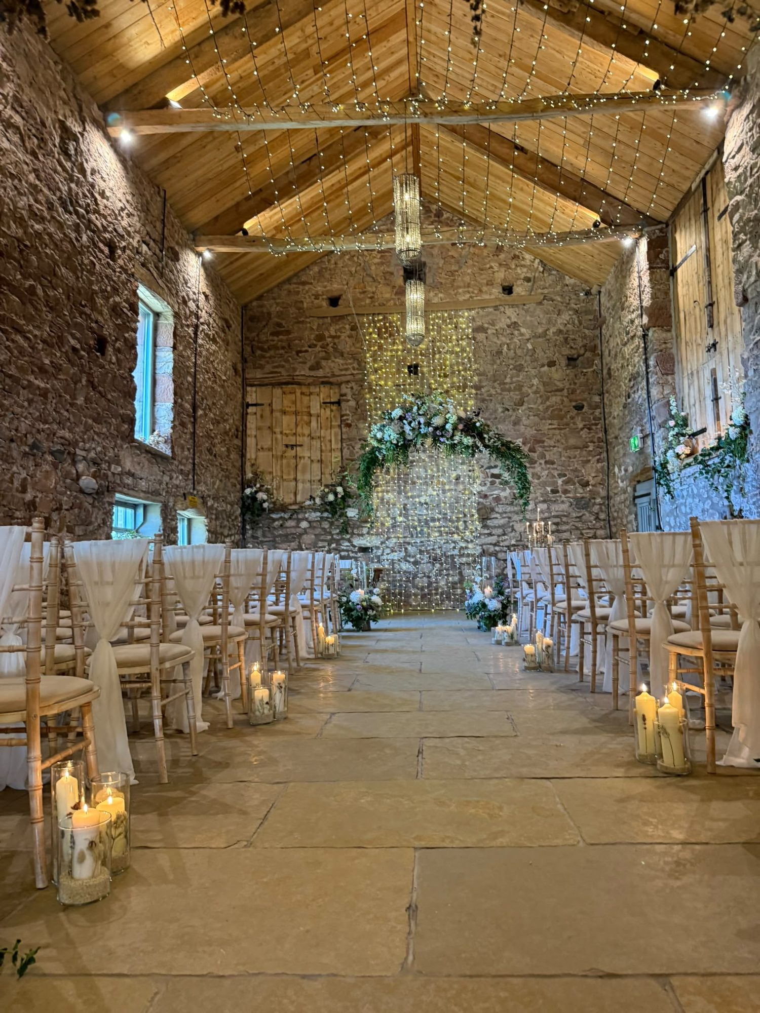A stone barn ceremony room dressed with a floral arch, fairy light curtain and candlelit aisle